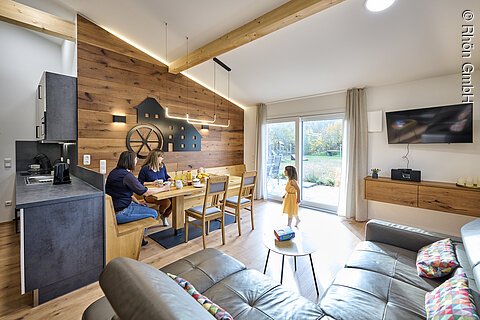Ein modernes Zimmer in Holzoptik, mit schwarzer Ledercouch, darin eine Familie am Tisch
